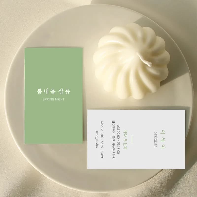 흰 접시 위 크림색 초 옆에 연두색 명함과 흰 명함을 놓은 사진